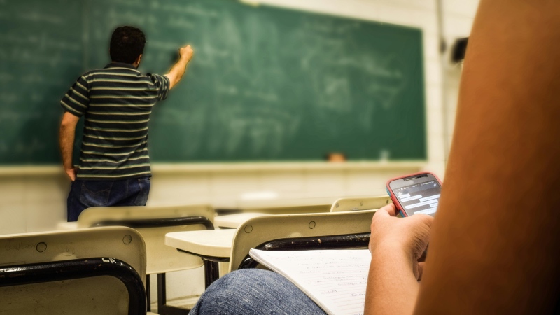 A student texts on their phone while a teacher writes on a blackboard in this undated stock image. (Pixabay/Pexels.com)