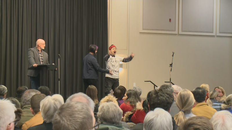 A man wearing a t-shirt that says "No tent in Ottawa" gets onto the stage at the Nepean Sportsplex during an information session about the  newcomer reception centre Sprung structure proposed for the area. Dec. 4, 2024. (CTV News Ottawa)