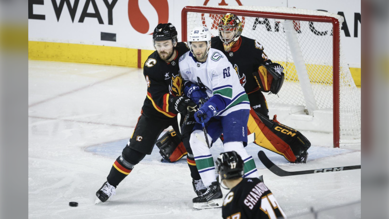 Vancouver Canucks' Max Sasson, centre, is checked by Calgary Flames' Rasmus Andersson, left, as goalie Dustin Wolf looks on during the first period of an NHL hockey game in Calgary on Tuesday, Dec. 31, 2024. THE CANADIAN PRESS/Jeff McIntosh