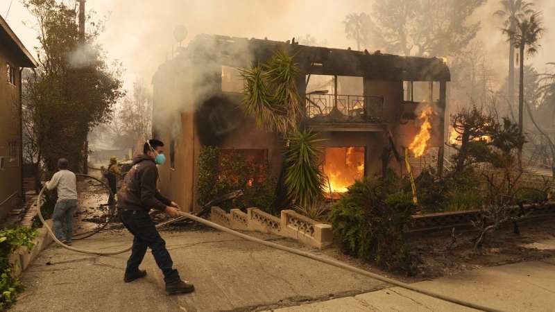 Pedestrians help a firefighter stretch a hose as an apartment building burns, Wednesday, Jan. 8, 2025, in the Altadena section of Pasadena, Calif. (Chris Pizzello / The Associated Press)